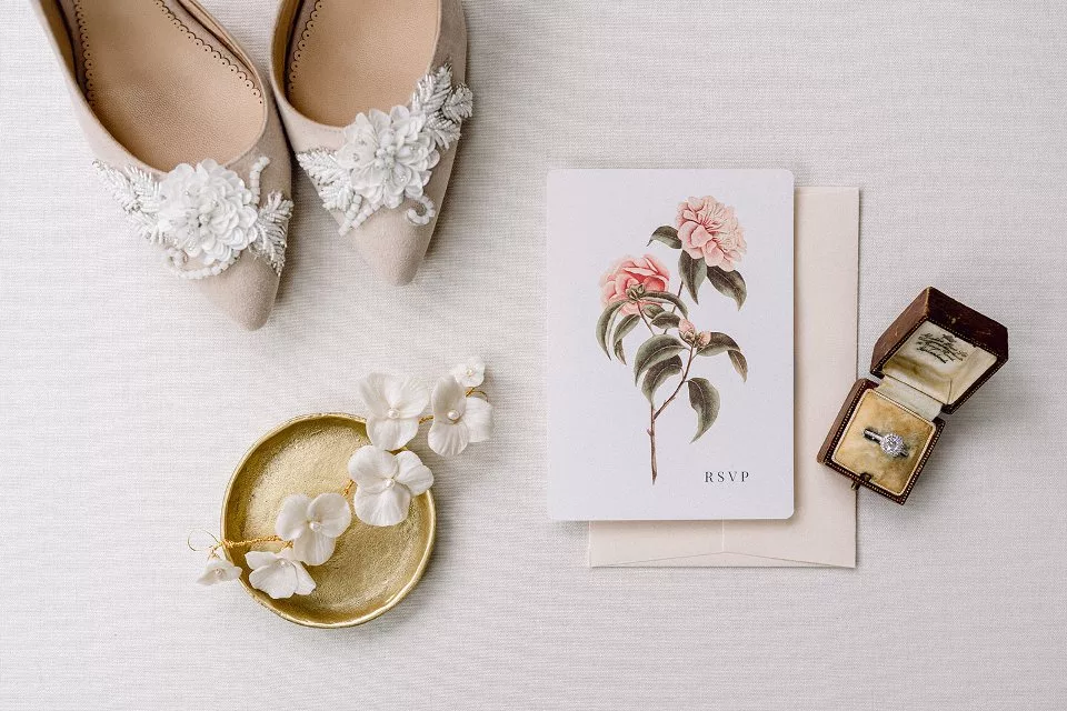 Flat lay beige background. A pair of shoes with floral trim near the toes sits in the top left corner. And gold jewellery tray sits below with a flower hair vine sitting in it. To the right is a card and envelope with a flower stem on, and to the right is an old jewellery box with engagement ring.