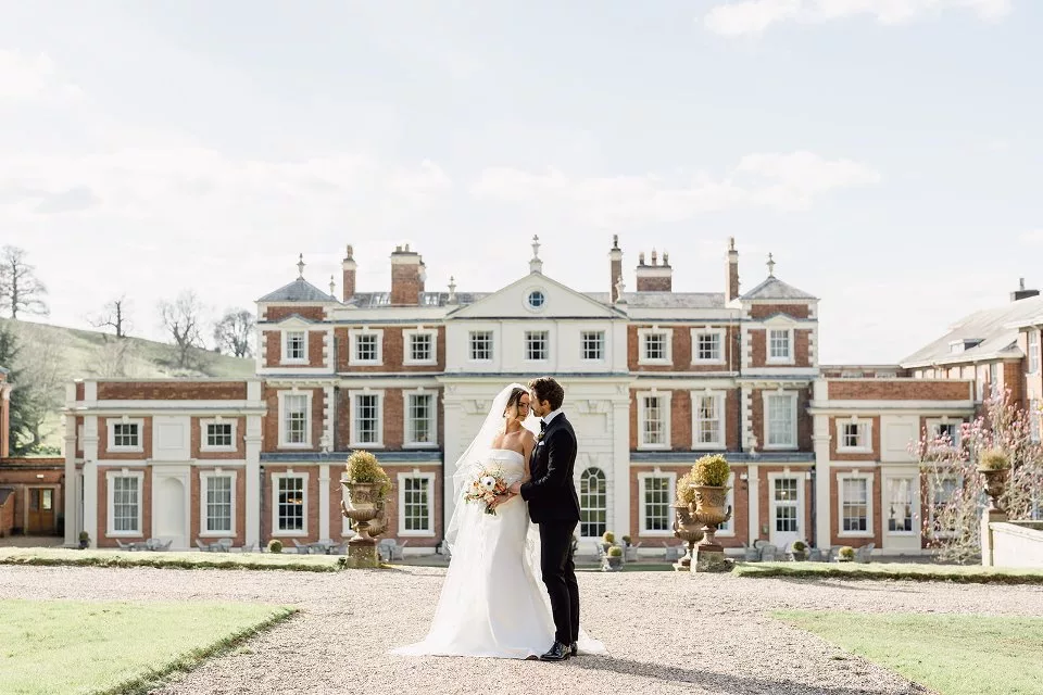 Bride and groom stand on the drive in front of a stately home.
