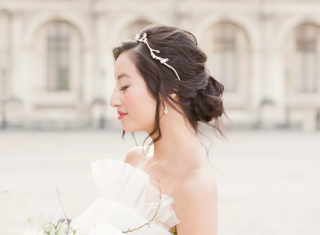 Bride stood in a Paris square with old buildings behind her. She is wearing a white gown and wears a Modern luxury romantic wedding hair accessory